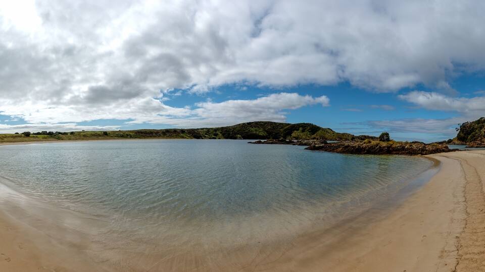 A beach with clear water, sandy shore, and green hills in the background.