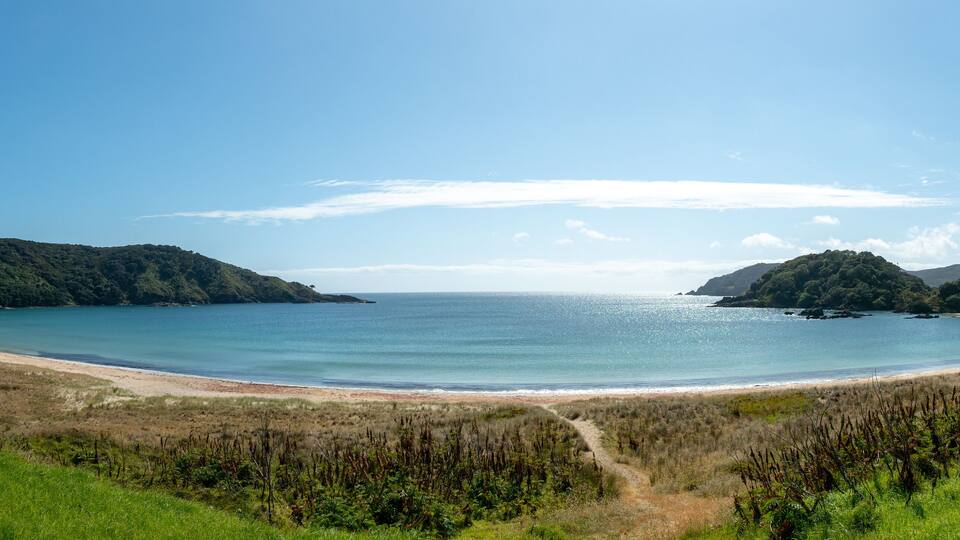 A coastal landscape with a sandy beach, a clear blue sea, and green hills in the background.