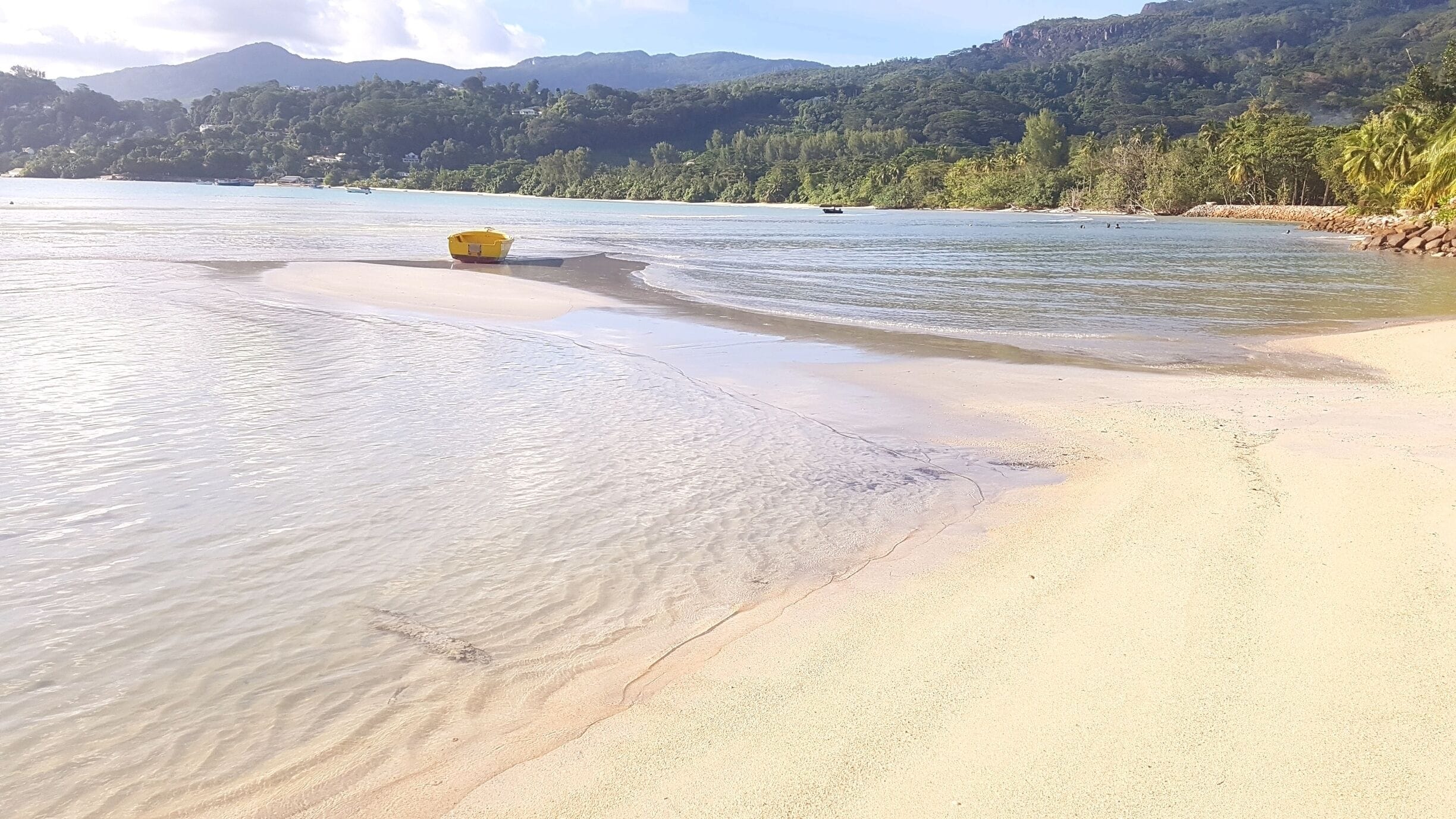 A beach with clear water, a yellow boat, and a forested hillside in the background.