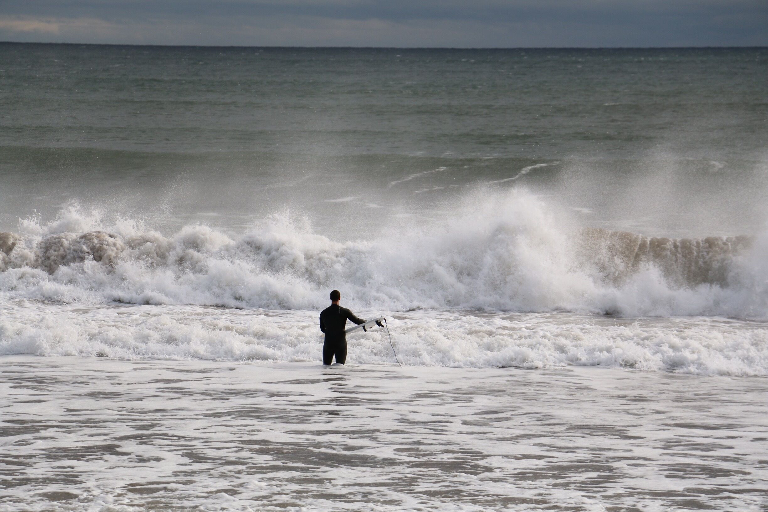 A person in a wetsuit fishing in the ocean with waves crashing nearby.