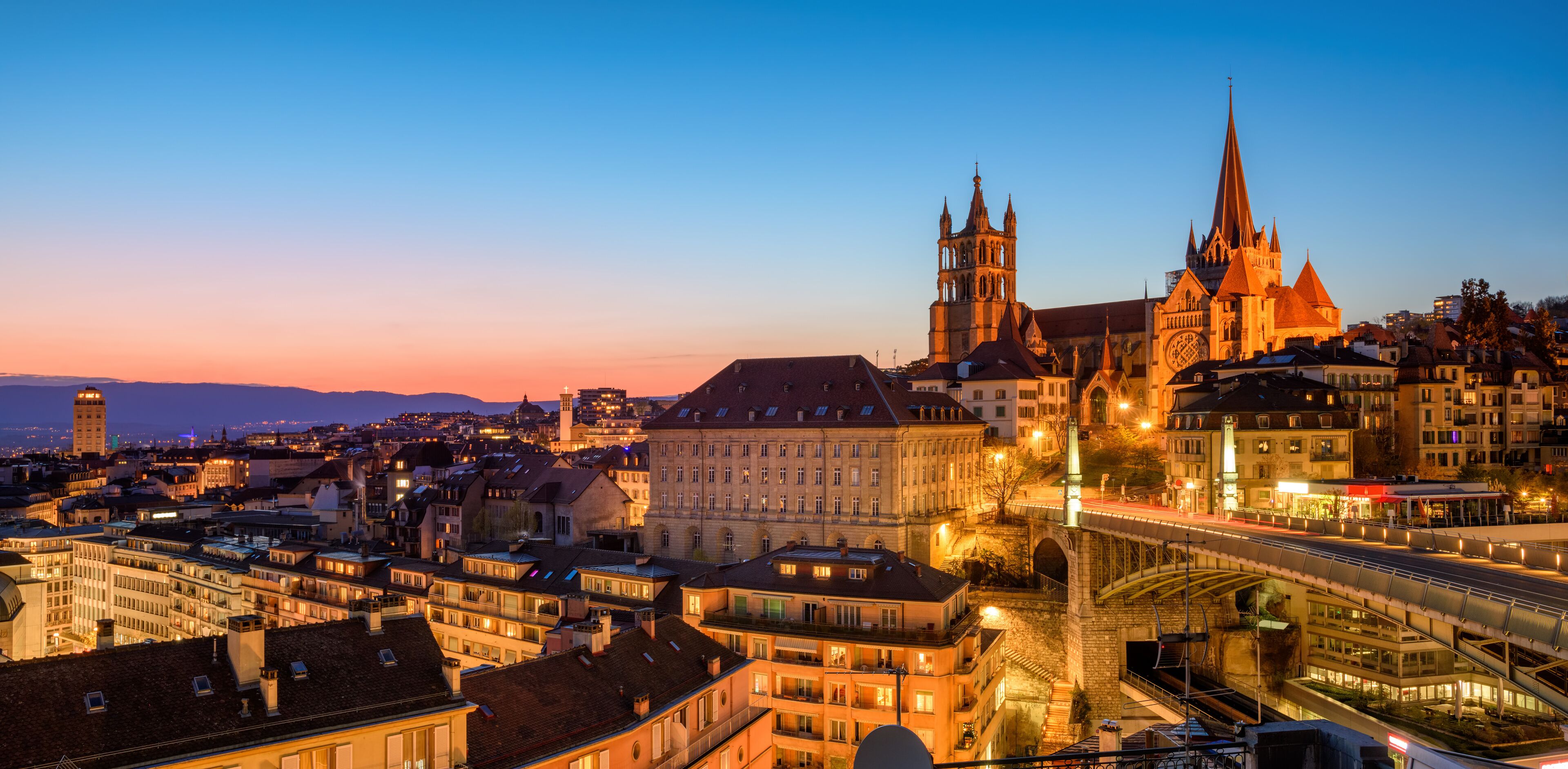 A cityscape at dusk with prominent Gothic architecture, including a cathedral with a tall spire and a bridge.
