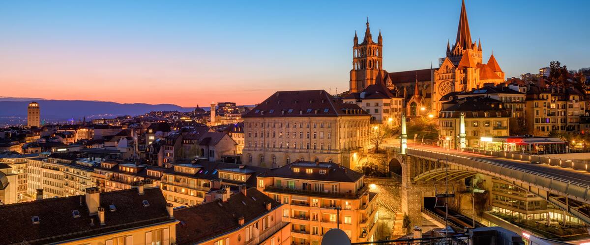A cityscape at dusk with prominent Gothic architecture, including a cathedral with a tall spire and a bridge.