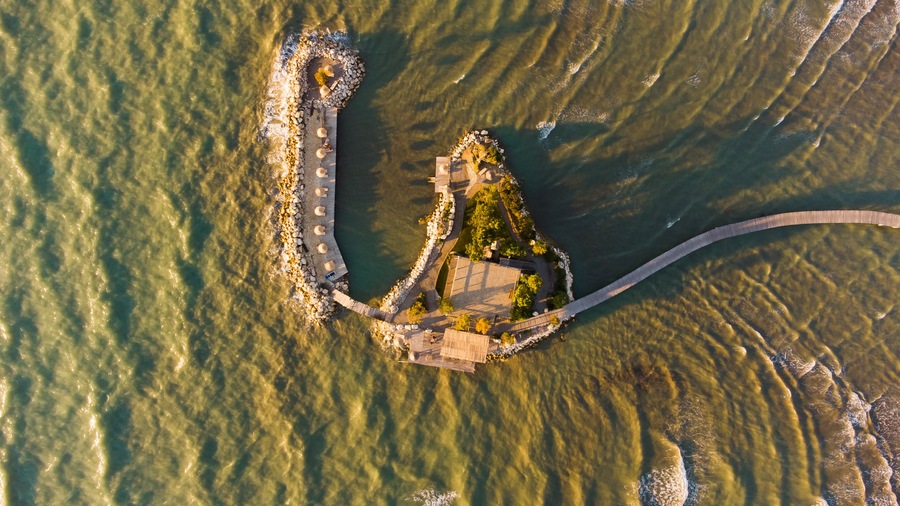Vista aérea de una zona costera con un muelle largo que se adentra en el agua, rodeado de rocas y vegetación.