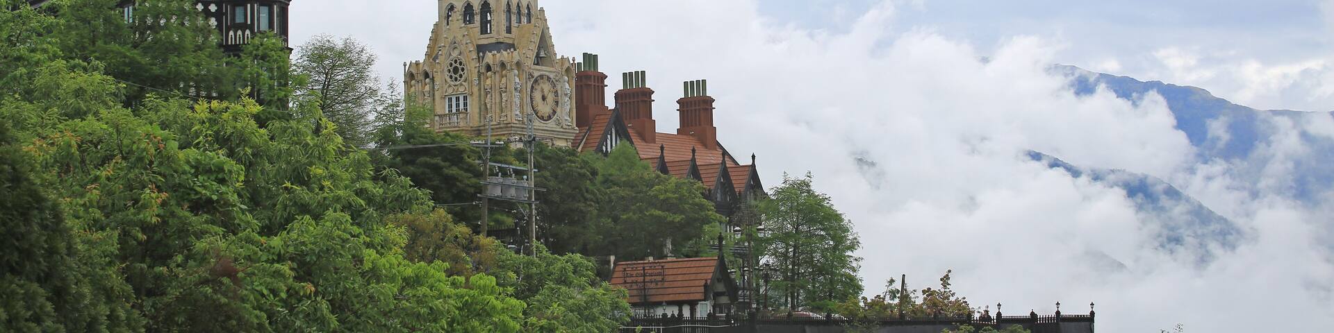A historic building with a clock tower and a church with a pointed roof, surrounded by lush greenery and clouds.