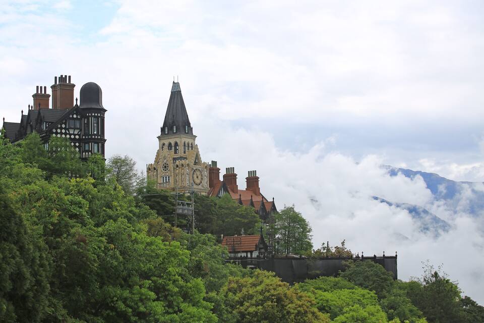 A historic building with a clock tower and a church with a pointed roof, surrounded by lush greenery and clouds.