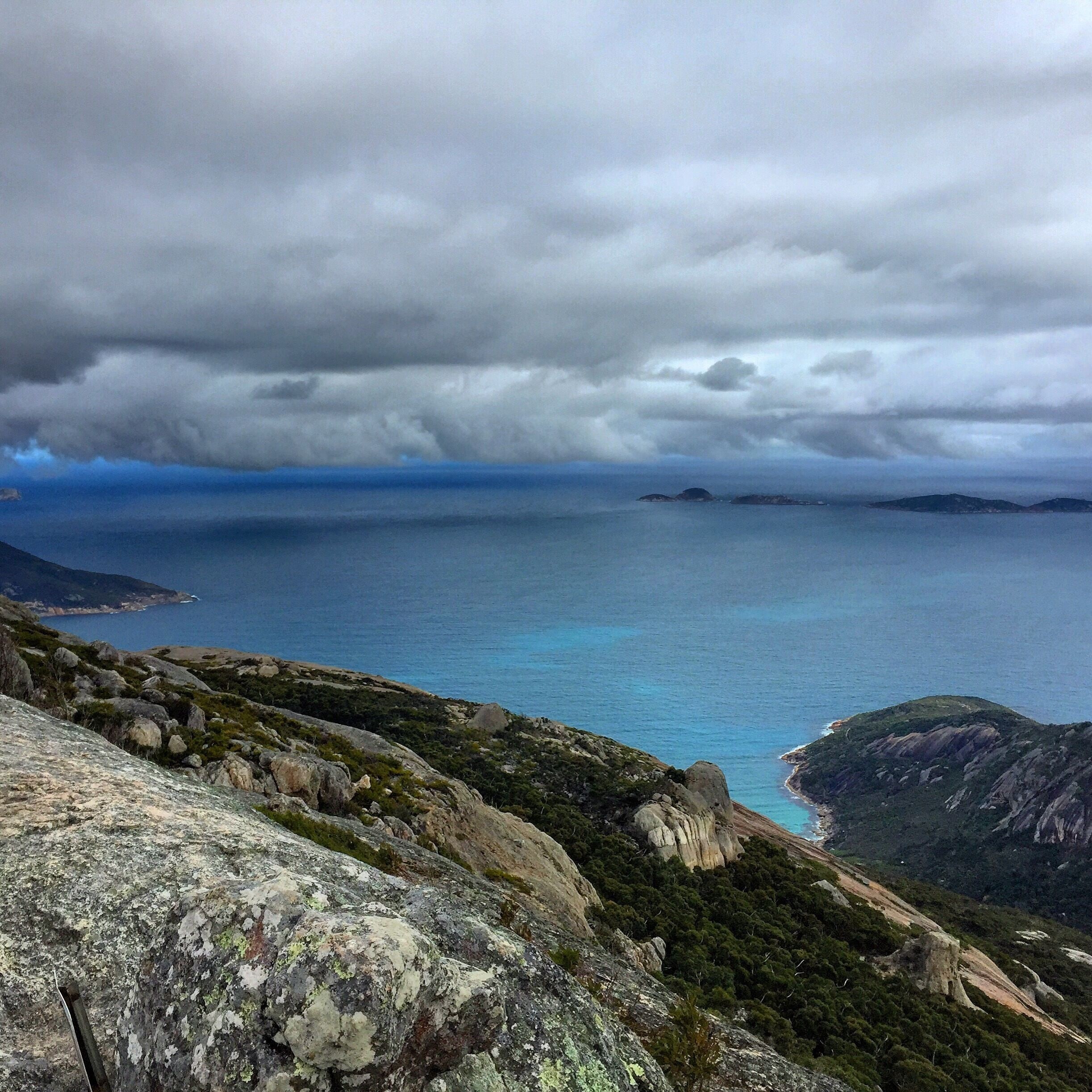 A coastal landscape with rocky terrain, a view of the sea, and distant islands under a cloudy sky.