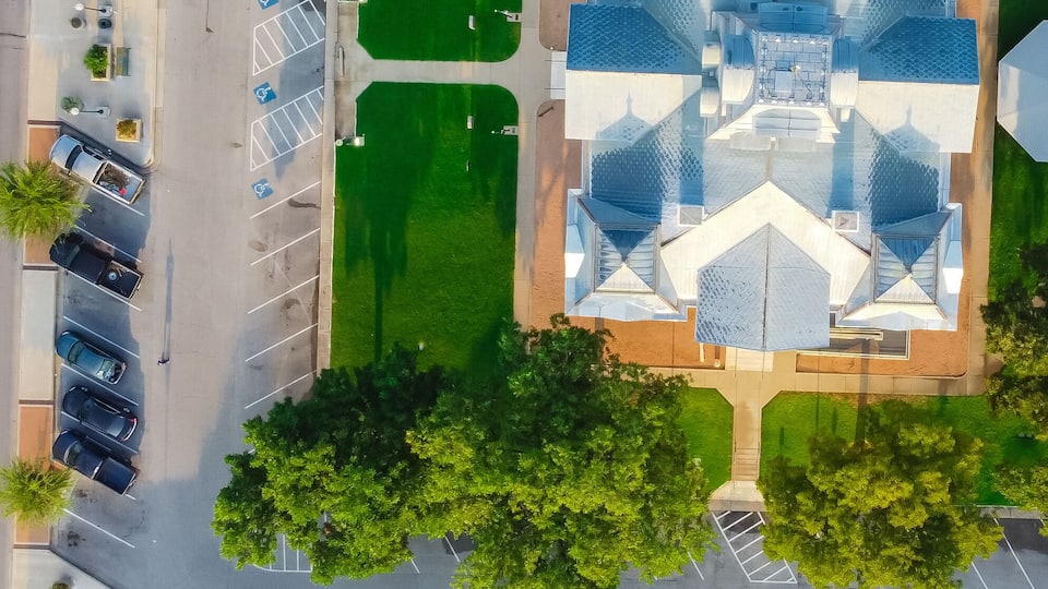 An aerial view of a city street with a central green park, trees, and a modern building with a unique roof design.