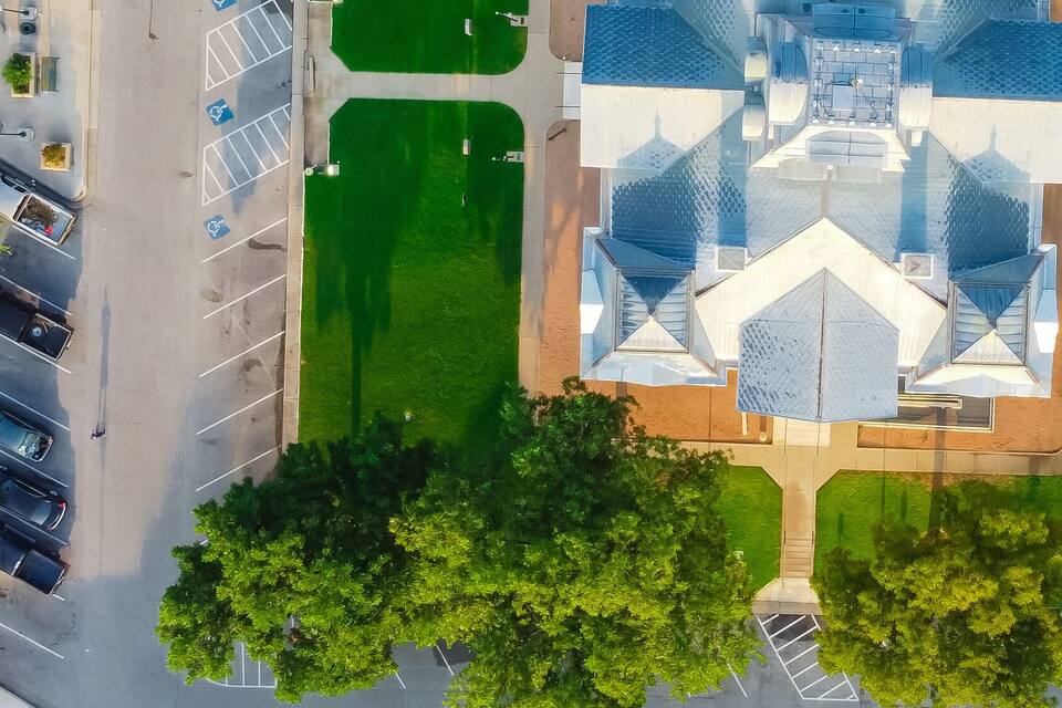 An aerial view of a city street with a central green park, trees, and a modern building with a unique roof design.