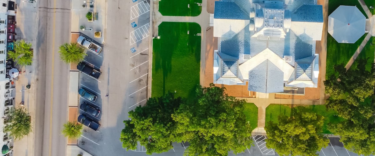 An aerial view of a city street with a central green park, trees, and a modern building with a unique roof design.