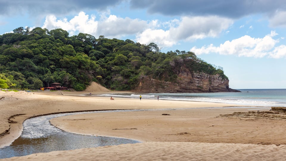 A sandy beach with a river flowing into the sea, a cliff with dense vegetation, and a partly cloudy sky.