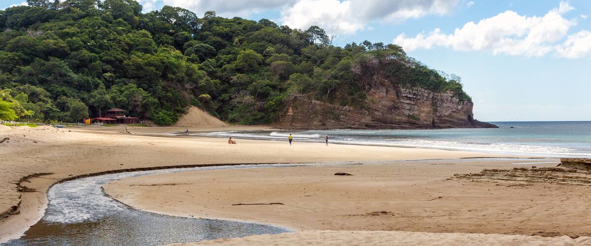 A sandy beach with a river flowing into the sea, a cliff with dense vegetation, and a partly cloudy sky.