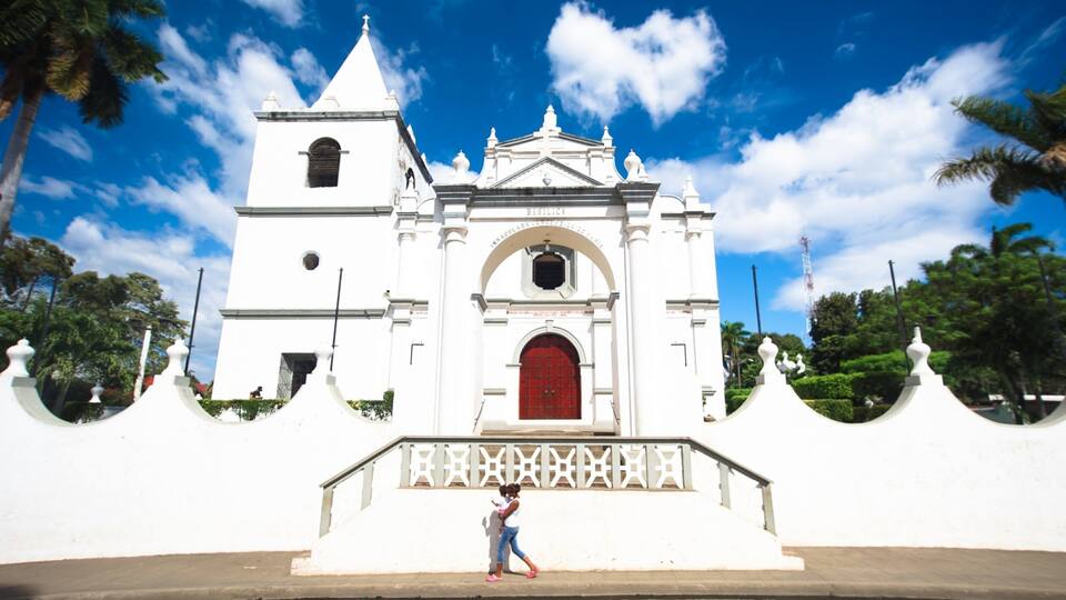 A white church with a red door and a bell tower, surrounded by white walls and palm trees.