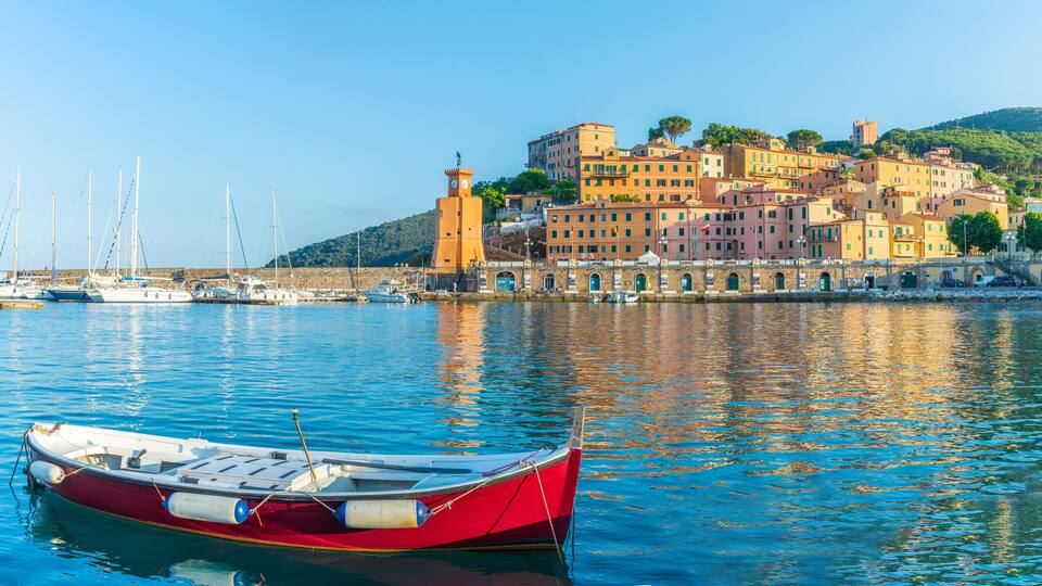 A red boat docked in calm waters, a historic tower, and colorful buildings on a hillside.