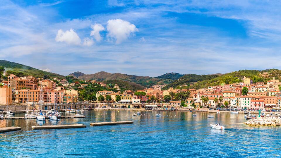 A coastal town with colorful buildings, a marina with boats, and hills in the background.