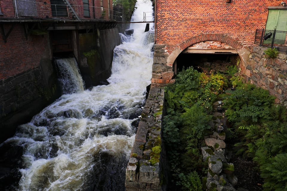 Une cascade d’eau dévalant un canal de pierre, entourée d’une végétation luxuriante et d’un bâtiment en briques doté d’une porte verte.