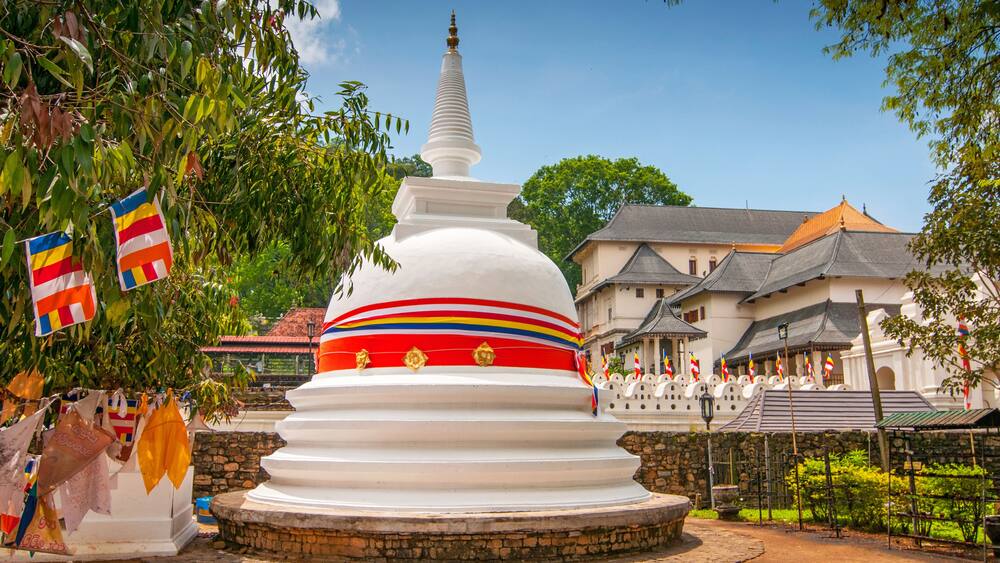 Un stupa blanc orné de bandes colorées, entouré de drapeaux et d’un bâtiment traditionnel.