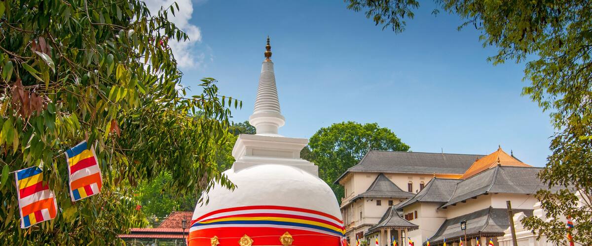 A white stupa with colorful stripes, surrounded by flags and a traditional building.