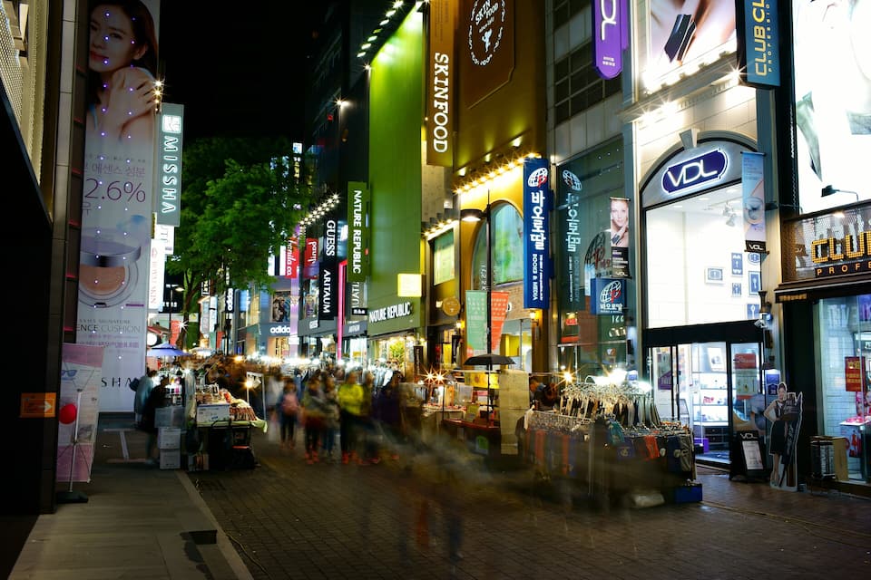 A bustling urban street at night with illuminated storefronts and a crowd of people.