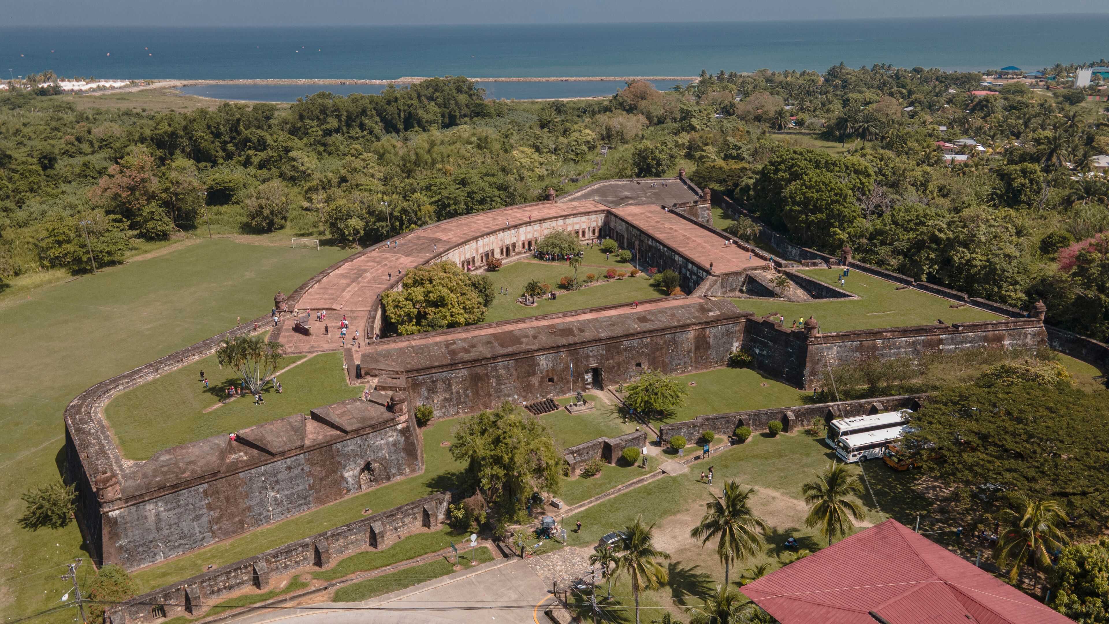 An aerial view of a historic fort with a curved wall, surrounded by lush greenery and palm trees, overlooking a coastal area.