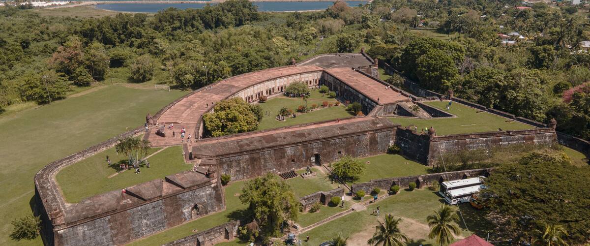 An aerial view of a historic fort with a curved wall, surrounded by lush greenery and palm trees, overlooking a coastal area.