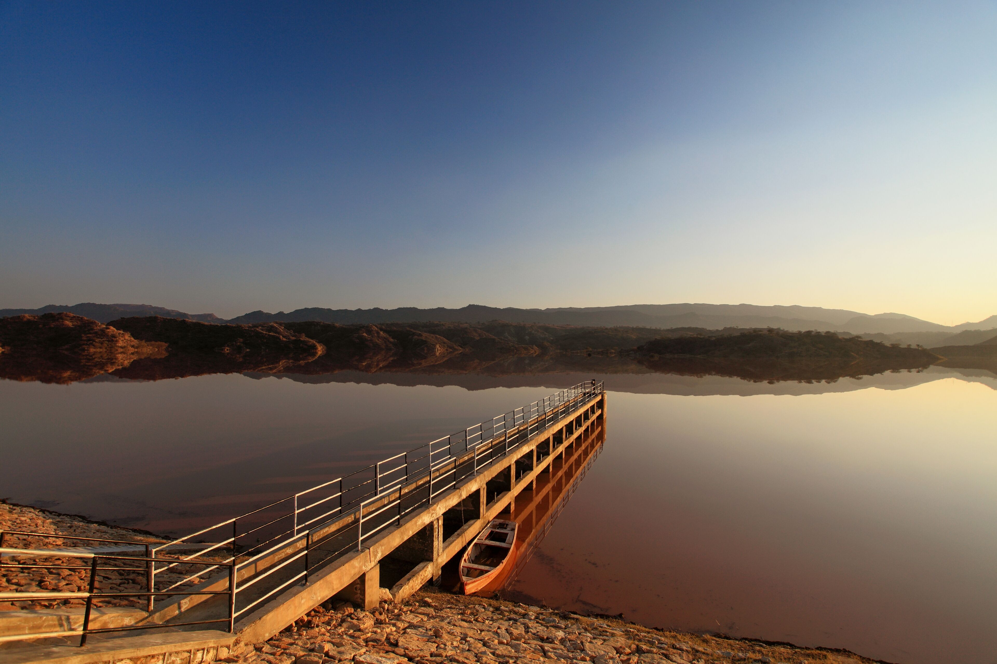 View of a long pier stretching into a tranquil lake reflecting the warm hues of the setting sun, with hills in the distance, Chakwal, Punjab, Pakistan.