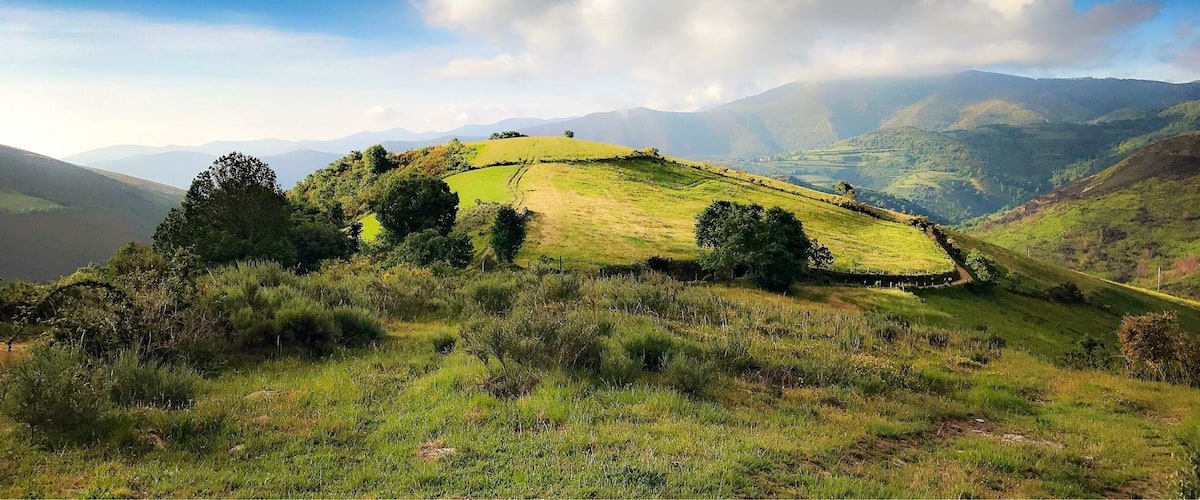 A grassy hillside with a fence, trees, and distant mountains under a partly cloudy sky.
