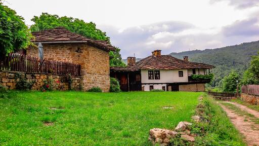 A rustic stone house with a tiled roof, surrounded by lush greenery and a stone wall.