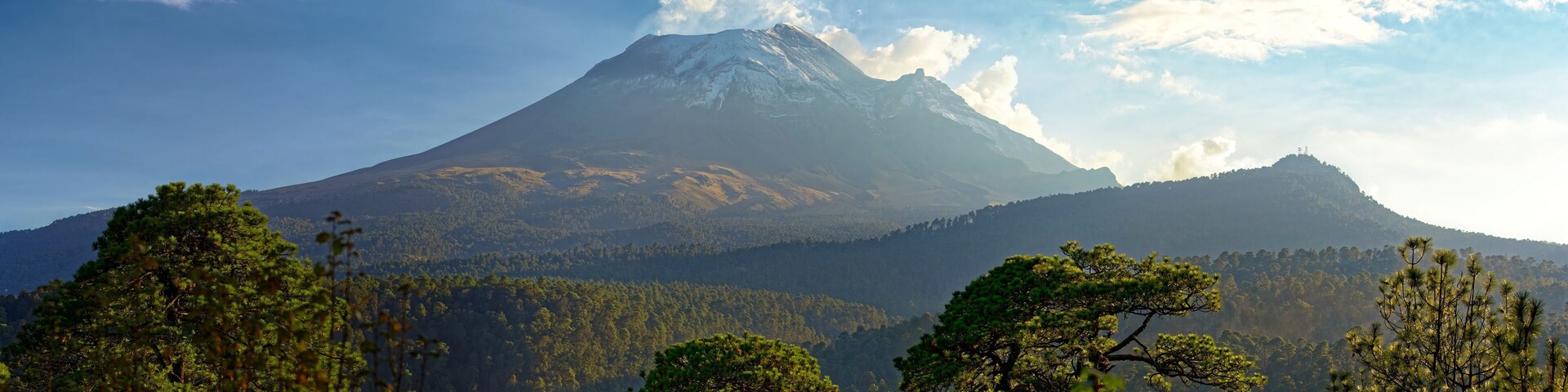 Popocatépetl volcano eruption smoke at the daytime among other green hills and mountains, with cloudy blue sky