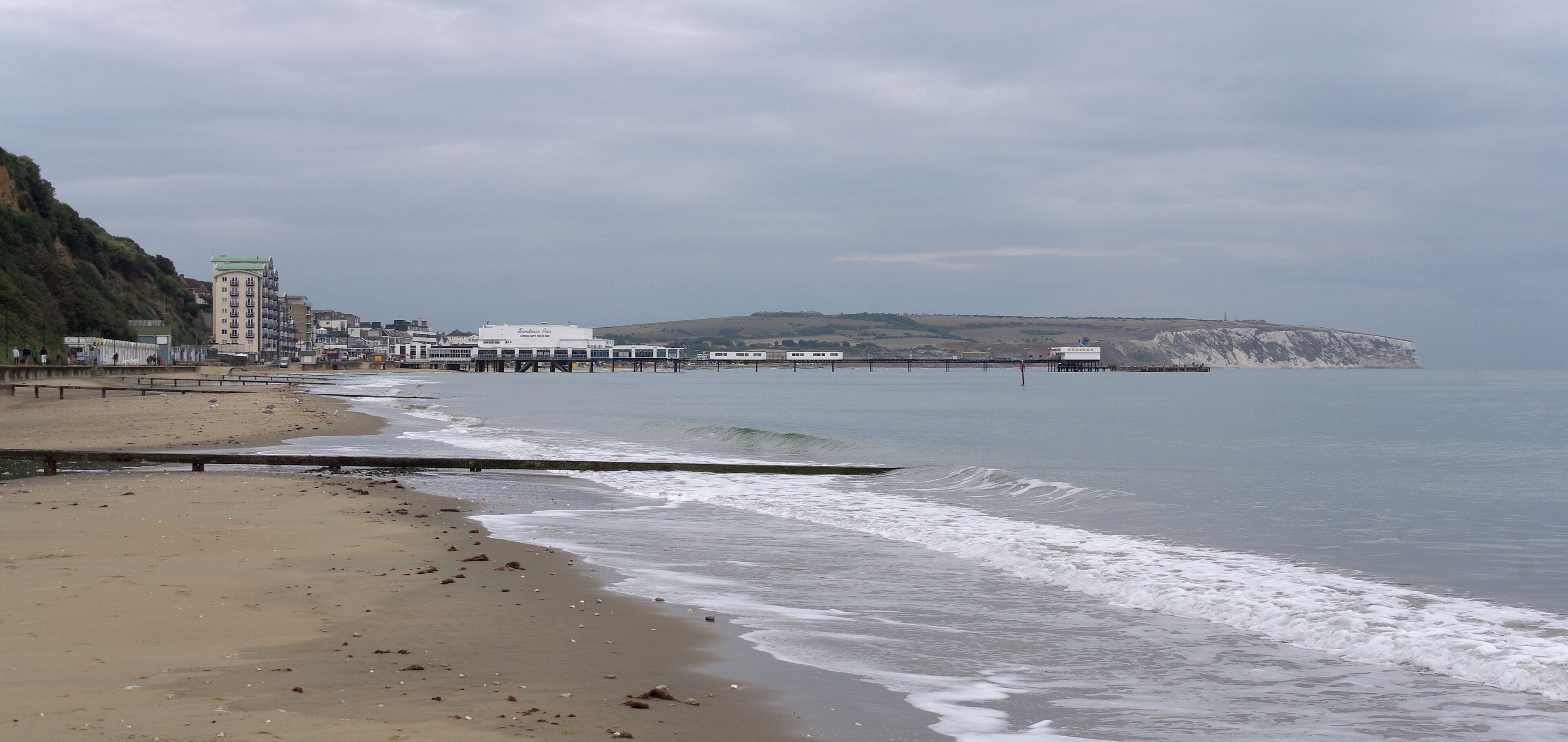 A beach with a pier extending into the water, buildings along the shore, and cliffs in the background.