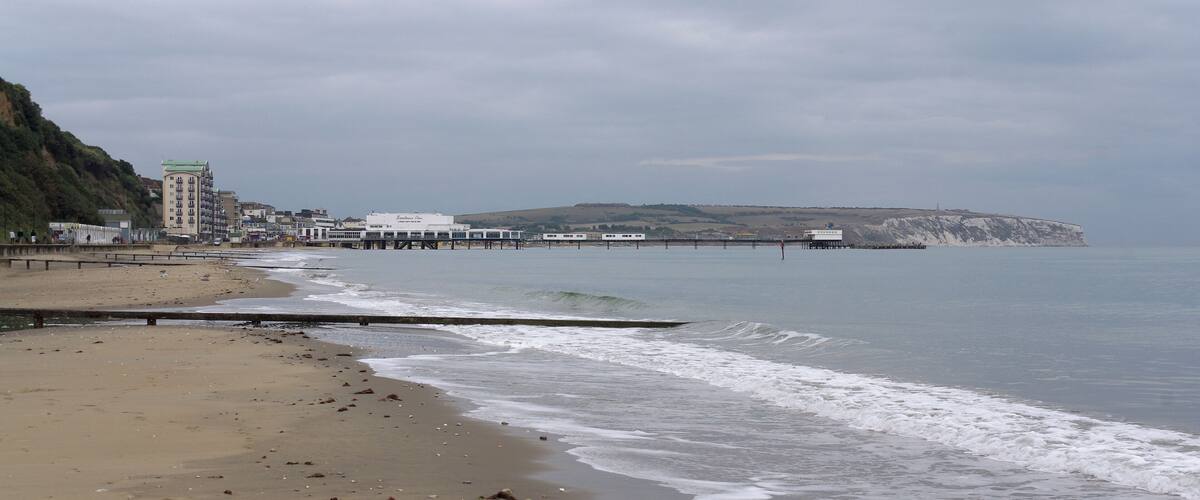 A beach with a pier extending into the water, buildings along the shore, and cliffs in the background.