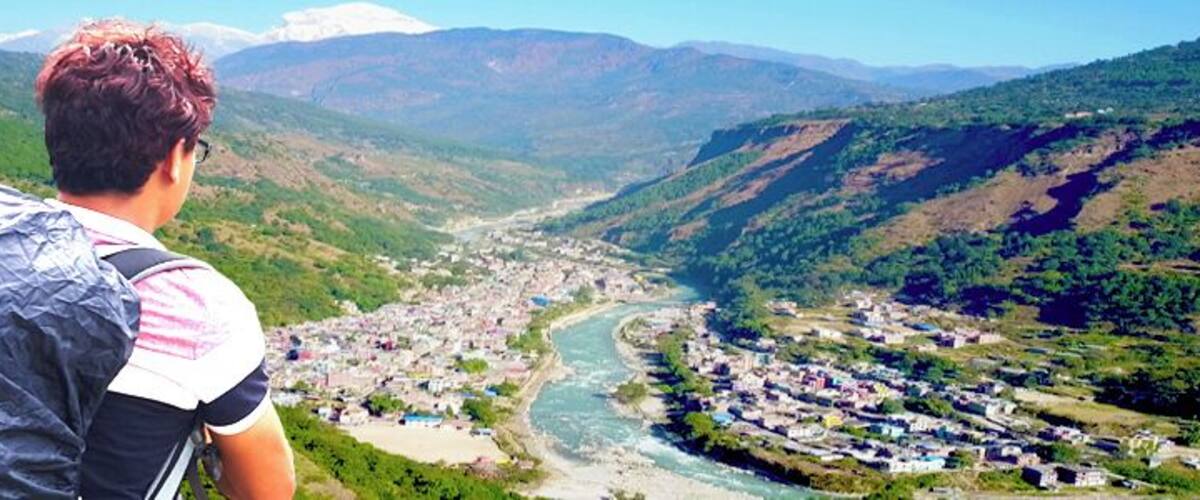 A person overlooking a valley with a river, surrounded by mountains and a small town.