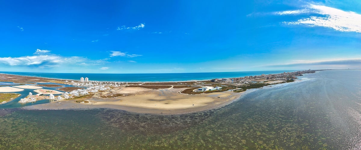 An aerial view of a coastal city with a beach, clear blue sky, and a distant city skyline.