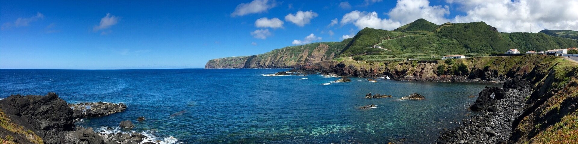 Paisagem costeira com costas rochosas, águas azul-claras e colinas verdes ao fundo.