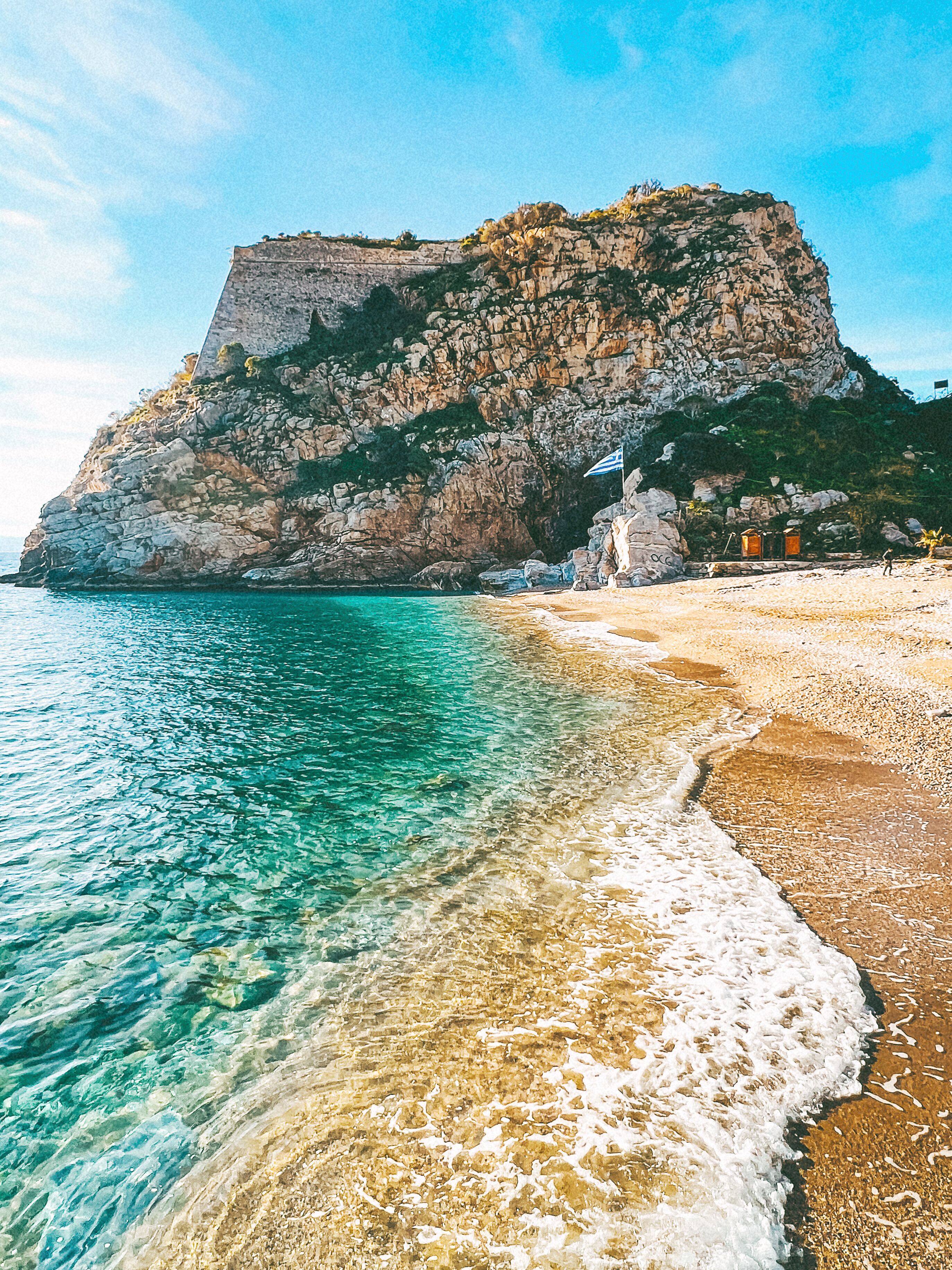 A beach with clear turquoise water, a rocky cliff, and a flag on top.