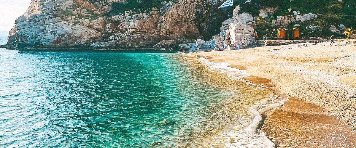 A beach with clear turquoise water, a rocky cliff, and a flag on top.