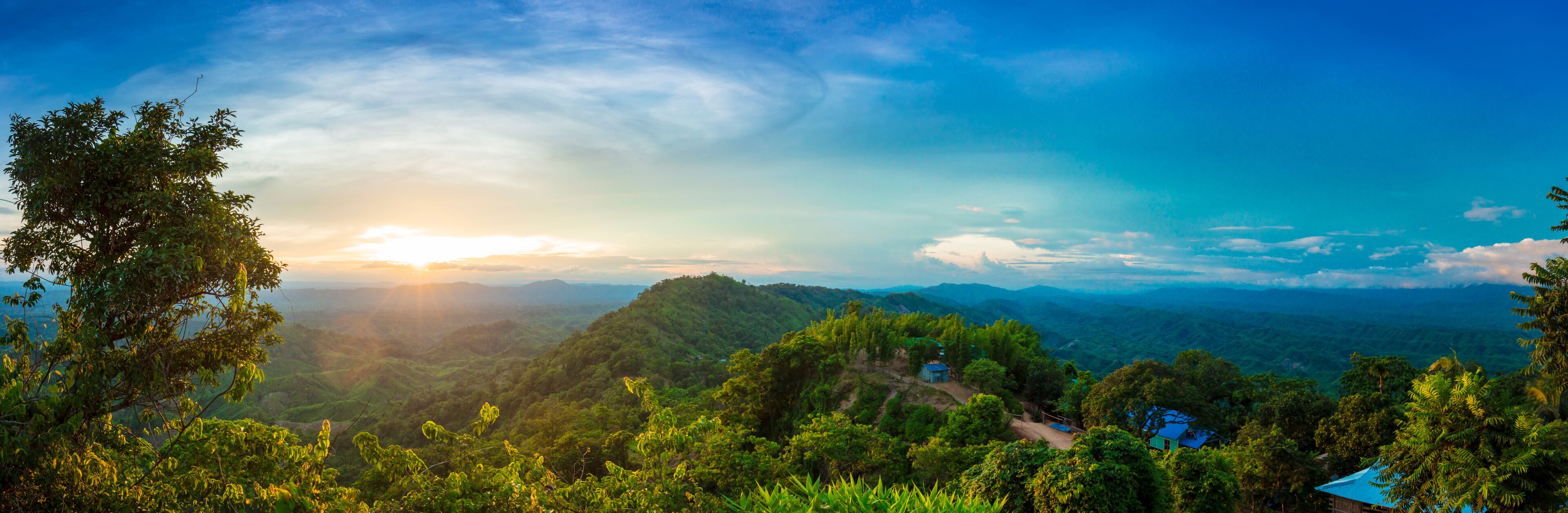 A lush green landscape with a clear sky, a sun setting or rising over a mountain range, and a small blue house in the foreground.