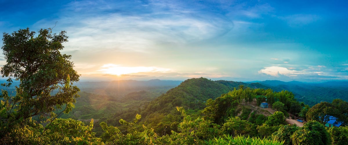 A lush green landscape with a clear sky, a sun setting or rising over a mountain range, and a small blue house in the foreground.