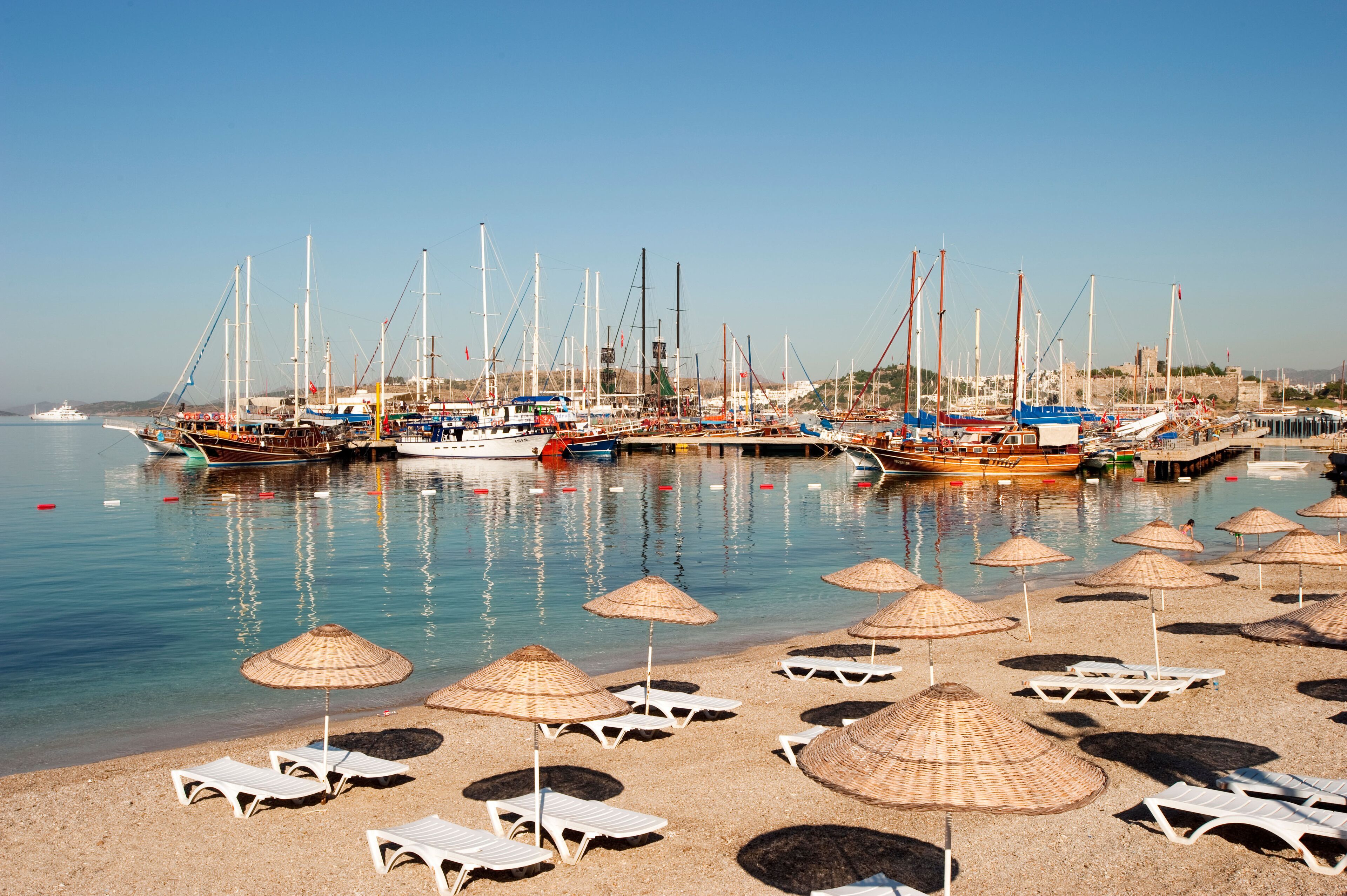 A marina with boats docked, a sandy beach with umbrellas, and a clear blue sky.