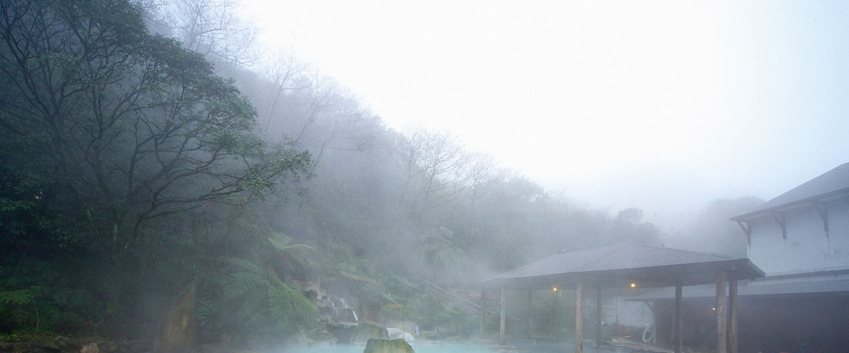 A hot spring surrounded by rocks and trees, with a building in the background.