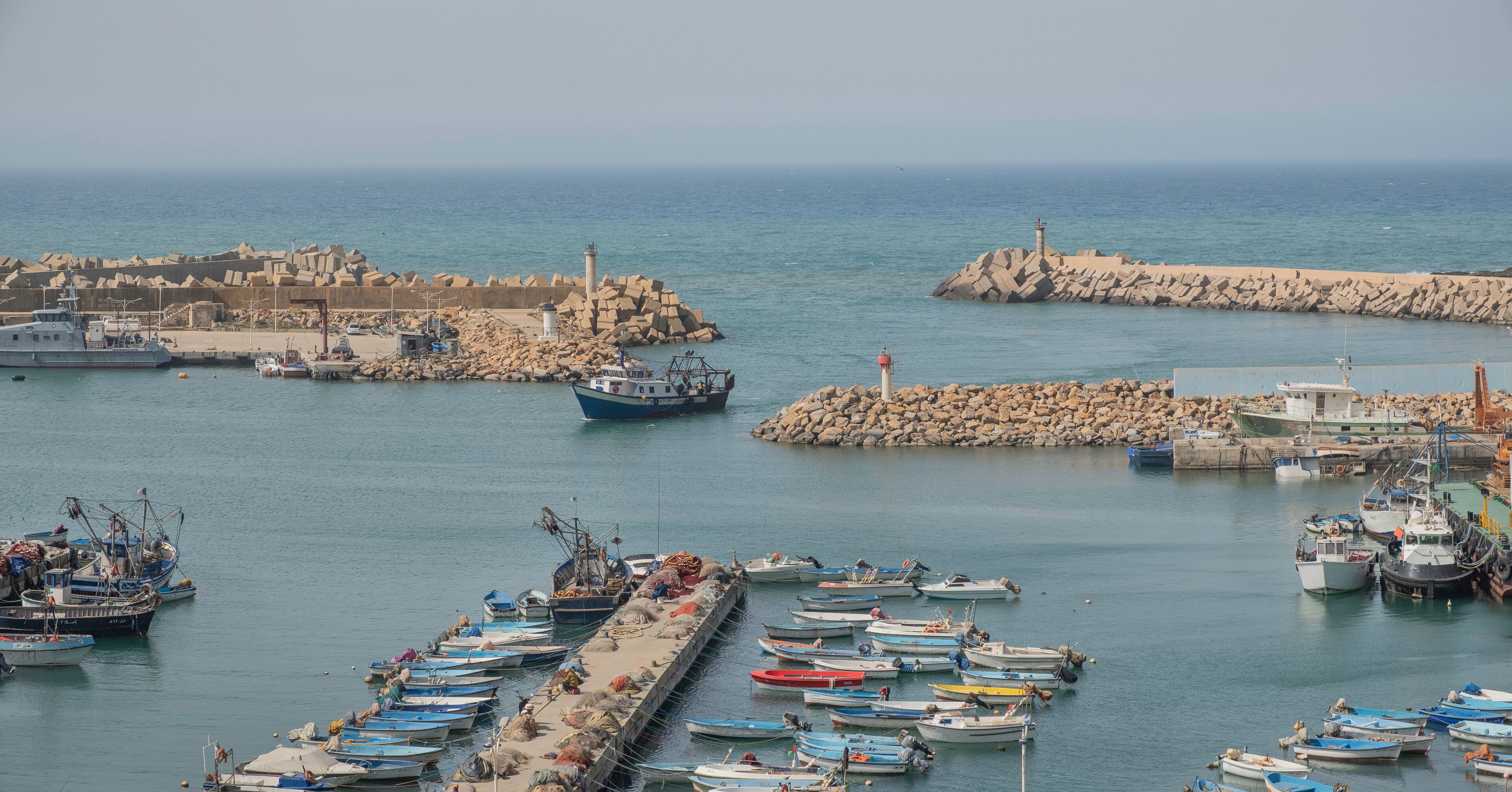 A harbor with numerous small boats docked, a lighthouse, and a rocky breakwater.