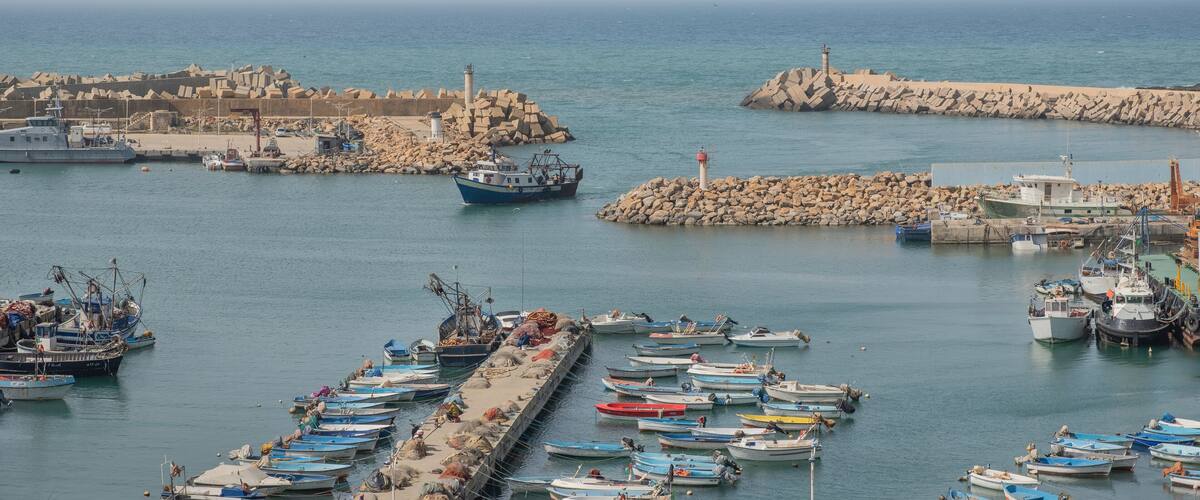 A harbor with numerous small boats docked, a lighthouse, and a rocky breakwater.