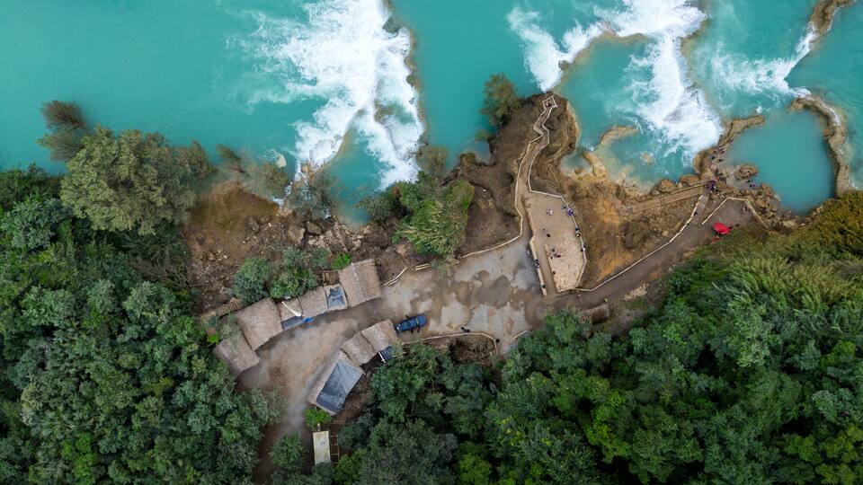 An aerial view of a coastal area with turquoise waters, rocky cliffs, and dense green vegetation.