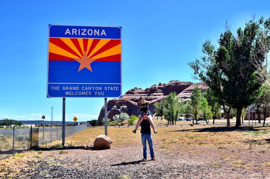 A person standing in front of a 'Welcome to Arizona' sign with a desert landscape in the background.