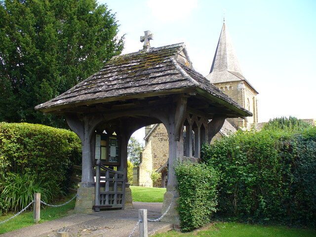 A stone gazebo with a pointed roof and a cross on top, surrounded by greenery.