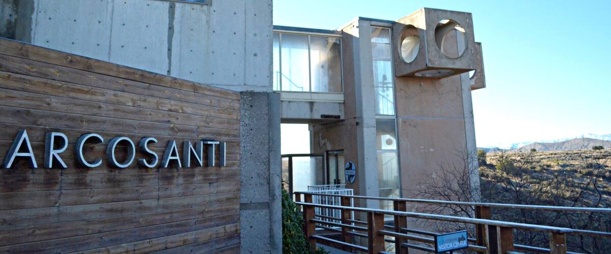 A building with the name 'ARCOSANTI' on a wooden wall, a modern architectural design with concrete and glass elements, and a mountainous landscape in the background.