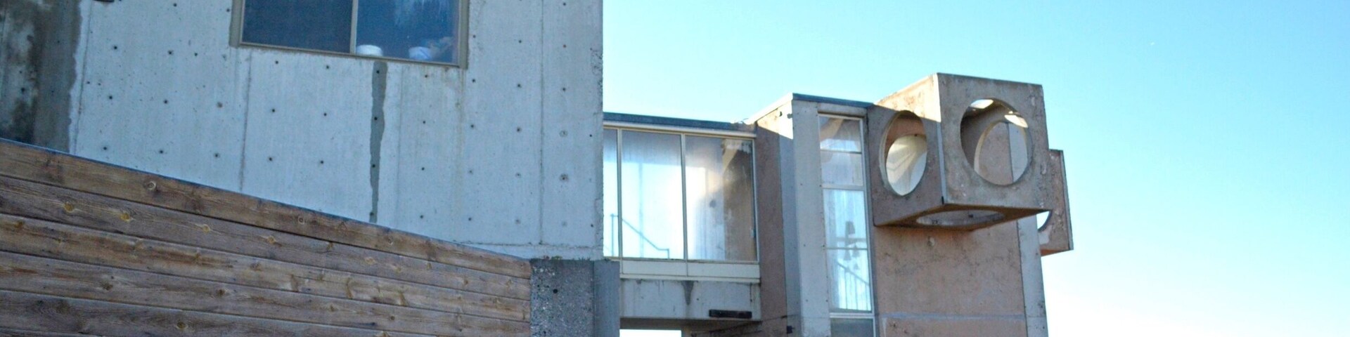 Um edifício com o nome 'ARCOSANTI' numa parede de madeira, design arquitetônico moderno com elementos de concreto e vidro, e uma paisagem montanhosa ao fundo.