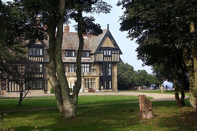 A large, historic house with a prominent tower and multiple windows, surrounded by a well-maintained lawn and trees.
