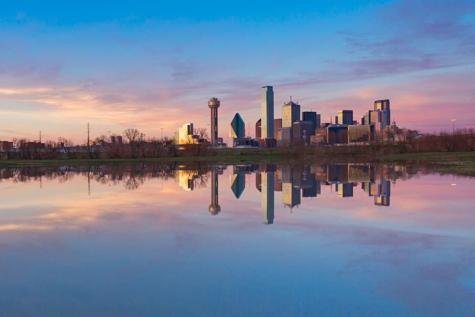 Lo skyline di una città riflesso in acque calme, un ponte e un cielo al tramonto.