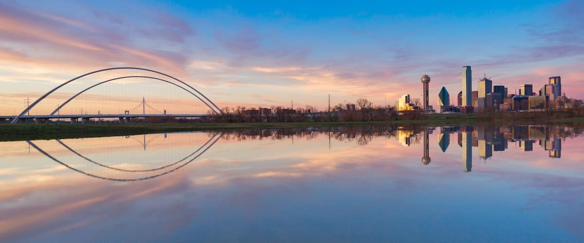 A city skyline reflected in a calm body of water, a bridge, and a sunset sky.