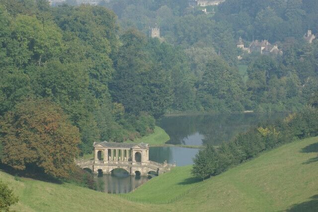 A tranquil lake with a gazebo-like structure on a bridge, surrounded by lush greenery and distant buildings.